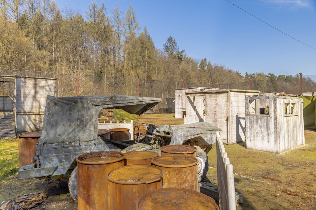 Auf einem Paintballfeld stehen ein altes Auto, rostíge Fässer und Holzhütten